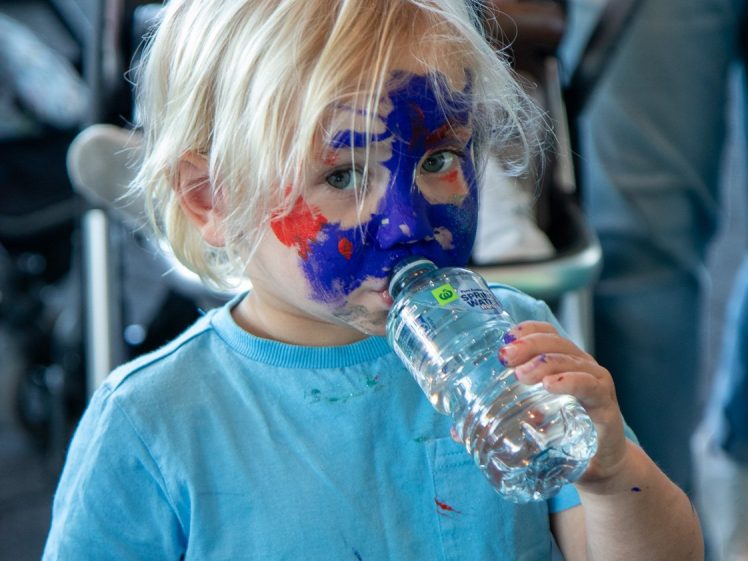 Image of A young child with blonde hair and colourful face paint drinks from a plastic water bottle. The child wears a light blue shirt, and the background shows blurred people and a pushchair.