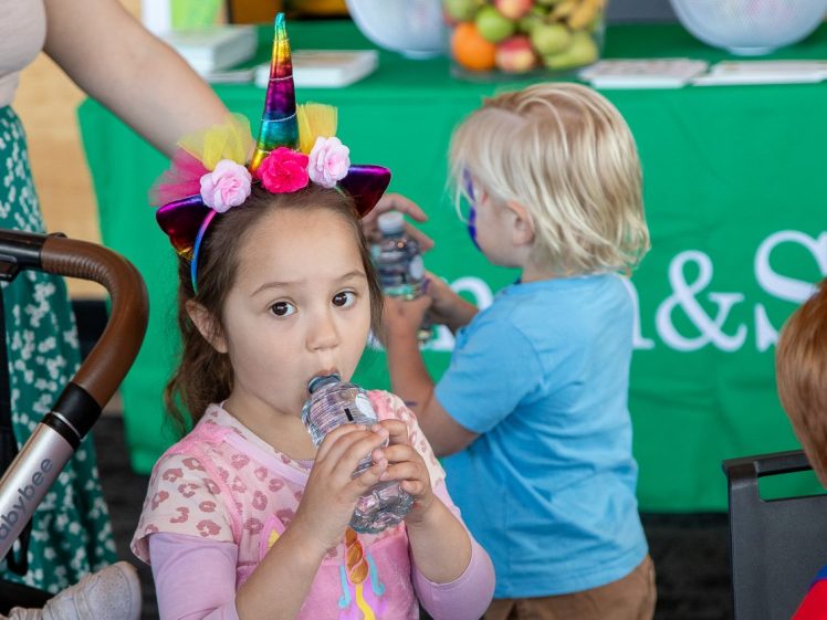 Image of A young girl wearing a colourful unicorn headband drinks from a water bottle. Behind her, a blonde boy holds a bottle. A table with bowls of fruit and a green tablecloth is in the background.