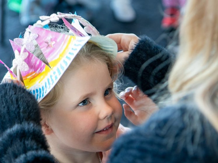 Image of An adult helps a smiling young girl put on a colourful, handmade paper crown decorated with stars and flowers. The scene suggests a fun, creative activity, likely at a party or craft event.