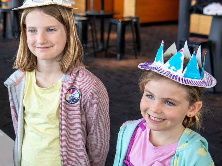 Image of Two young girls wearing handmade paper crowns with colourful decorations, smiling indoors. One girl has a striped jacket and yellow shirt, the other a light blue jacket and purple dress. The background is a modern room with tables and stools.
