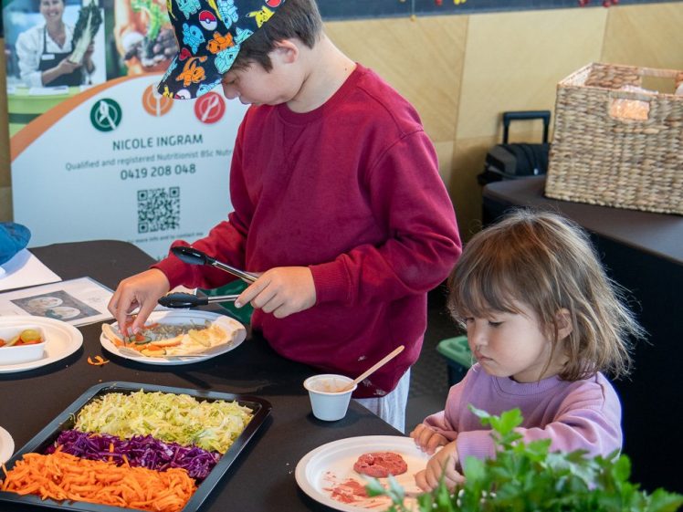 Image of Two young children sit at a table preparing their own meals with fresh vegetables and other ingredients. Colourful chopped veg and herbs are on the table. Workshop flyers are displayed in the background.