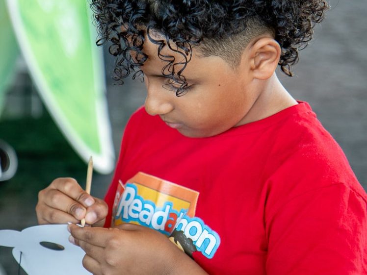 Image of A child with curly hair wearing a red Readathon T-shirt is focused on making a craft, threading a string through a white paper mask.