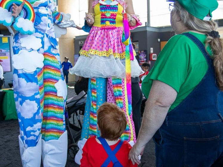 Image of Two people on stilts dressed in colourful rainbow costumes and wigs smile at a child in a Mario costume, while an adult in green and blue dungarees stands nearby. The setting appears to be an indoor event.