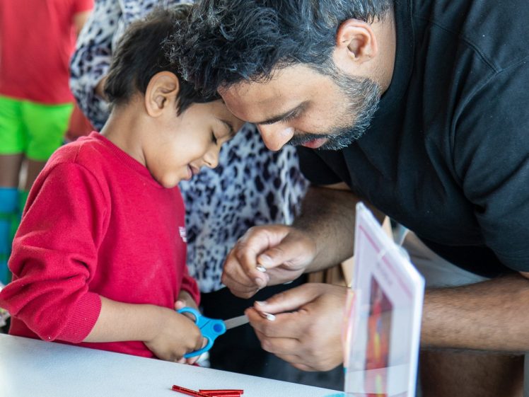Image of An adult helps a smiling child with a craft activity at a table. The child holds scissors, and colourful craft materials are scattered on the table. Other people are visible in the background.