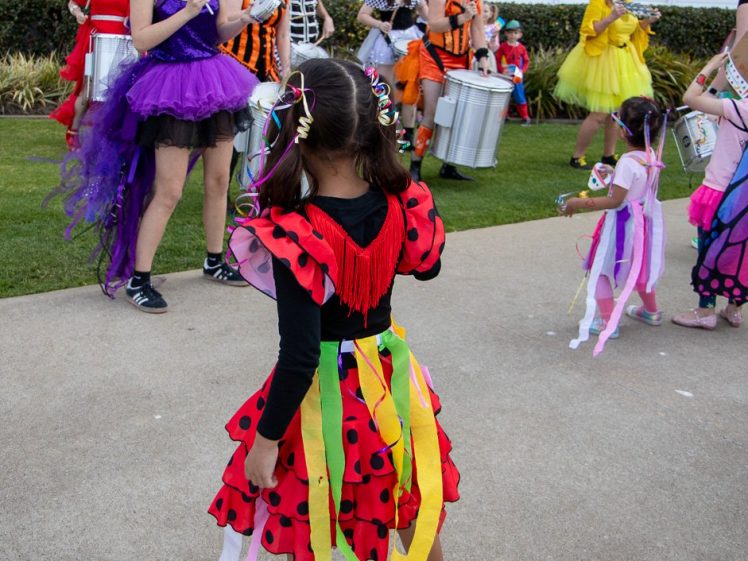 Image of A young girl in a red ladybird costume with colourful ribbons watches a group of adults and children dressed in vibrant, whimsical costumes playing drums outdoors near a white building.