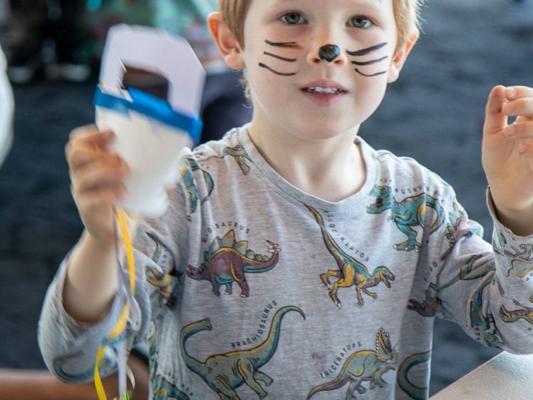 Image of A young child with whiskers and a nose drawn on their face, wearing a dinosaur-patterned shirt, holds colourful ribbons and smiles whilst sitting at a table.