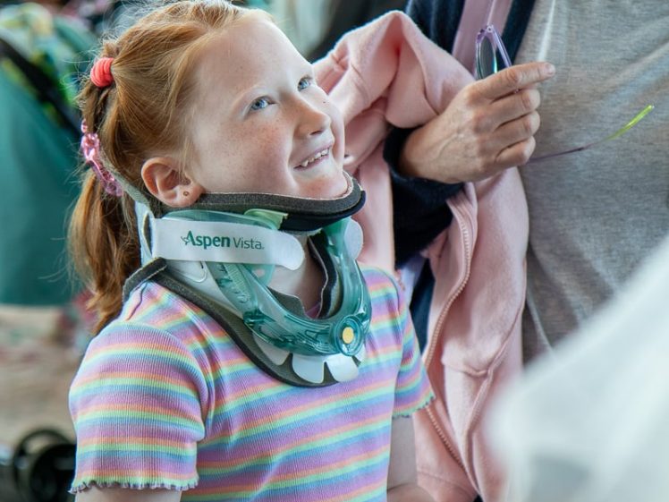 Image of A smiling young girl with red hair and a striped shirt wears a neck brace and stands beside an adult woman holding a pink jacket. People are visible in the blurred background.