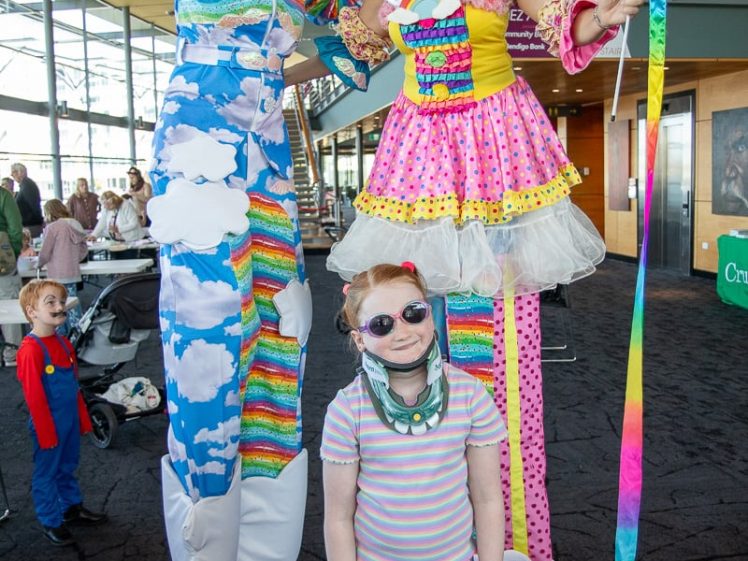 Image of Two adults in colourful, cloud- and rainbow-themed costumes on stilts pose with a smiling child in sunglasses and floral clothes. Other people are in the background, including a child in a Mario costume. Indoors, festive atmosphere.