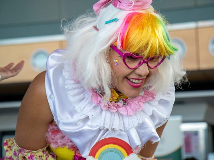 Image of A person dressed as a colourful clown with rainbow hair, heart-shaped pink glasses, white ruffled collar, and a rainbow prop, smiling and leaning forward indoors.