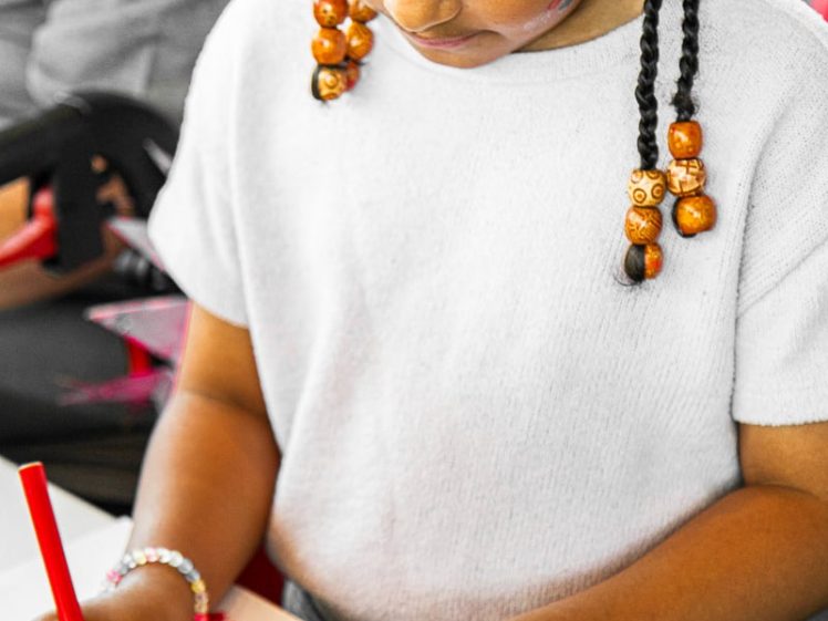 Image of A young girl with braided hair and beads colours red circles on paper with a red felt-tip pen whilst sitting at a table, focused on her drawing activity.