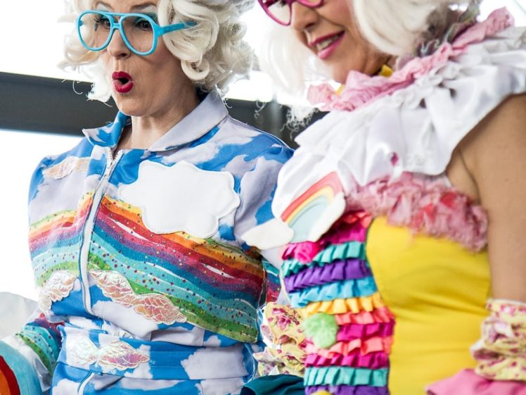 Image of Two people wearing colourful, whimsical costumes with cloud and rainbow motifs, puffy wigs, and bright glasses, stand together indoors, appearing cheerful and animated.