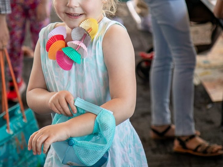 Image of A young girl with blonde hair in a light blue dress and colourful necklace smiles while holding a small blue handbag. Other people stand and walk in the background of an indoor setting.