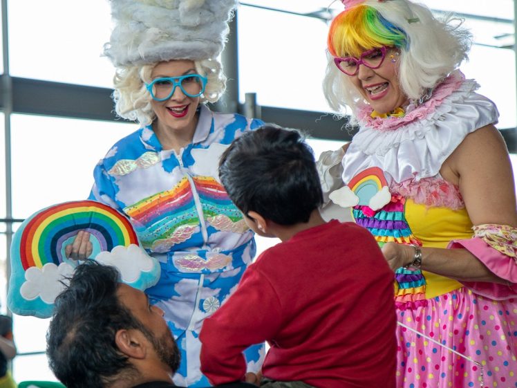 Image of Two people in colourful costumes and wigs, holding rainbow props, smile and interact playfully with a child and an adult in a bright indoor setting.