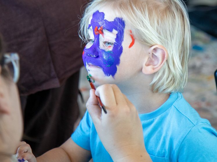 Image of A young child with blonde hair and a blue shirt gets their face painted with bright purple, green, and red colours by an adult holding a brush. The child looks ahead whilst the artist works.