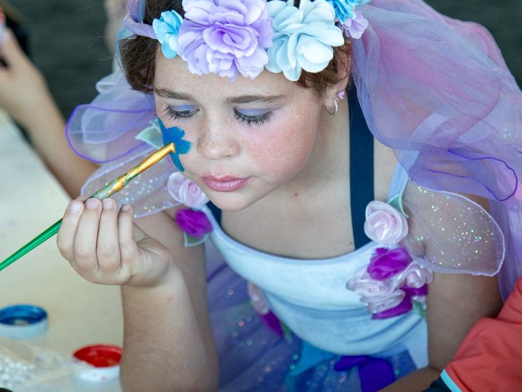 Image of A young girl wearing a flower crown and a colourful costume sits at a table while having her face painted with a blue star. Other children and painting supplies are visible around her.