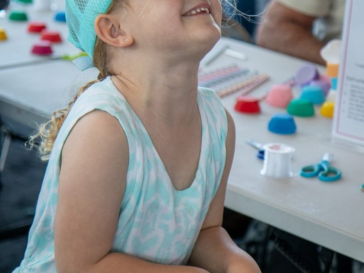Image of A young girl in a blue dress and matching cap smiles and looks up whilst standing near a table with colourful craft supplies and scissors at an indoor activity area.