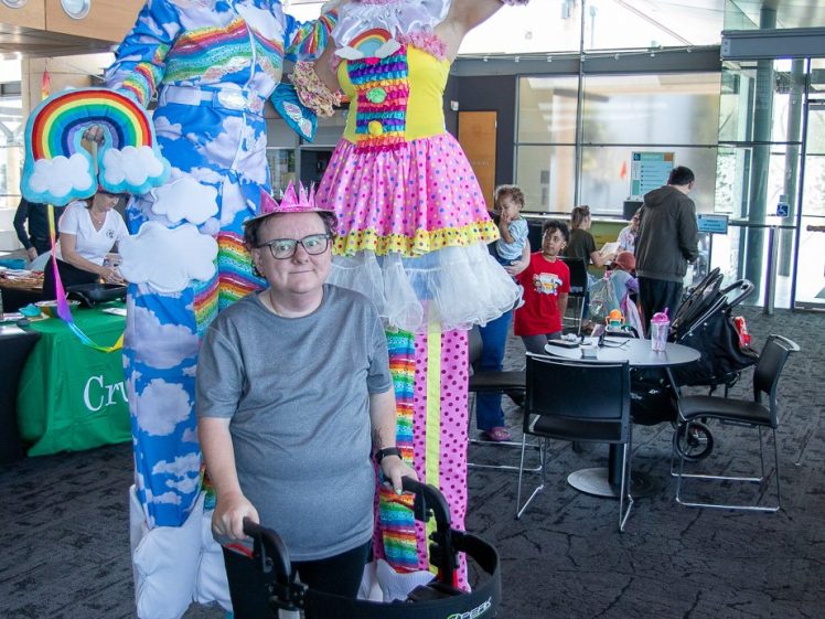 Image of Two people on stilts dressed in colourful, whimsical cloud and rainbow costumes pose with a smiling person using a walking frame. There are tables and people in the background at an indoor event.