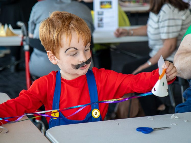 Image of A young child dressed as Mario from Super Mario examines a colourful ribbon and a decorated paper craft at a table during a craft activity. Other people are seated and working in the background.