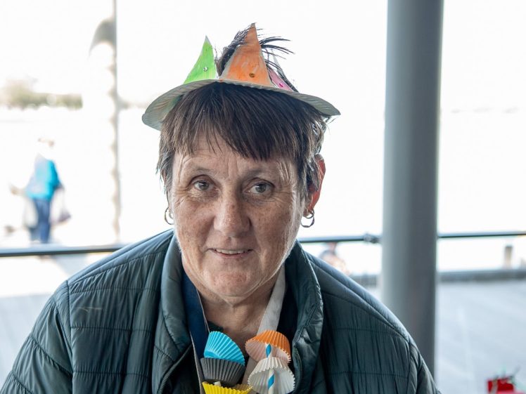 Image of A woman wearing a homemade paper hat and a jacket smiles at the camera. She also has colourful paper cupcake cases clipped to her shirt. The background is bright and slightly out of focus.