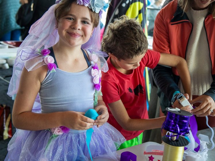 Image of A smiling child in a purple fairy costume and flower crown stands at a craft table with ribbons, while another child and an adult work beside them. The setting appears lively and festive, with people in the background.