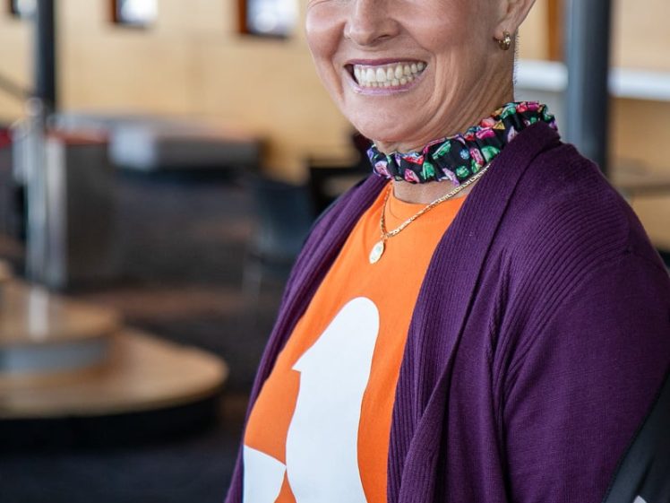 Image of Smiling woman with short grey hair wears a colourful floral crown, a purple cardigan, and an orange shirt with a white logo. She also wears a pink skirt and stands indoors with blurred background.