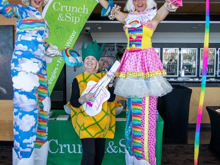 Image of Two people on stilts dressed in colourful, whimsical costumes stand on either side of a person in a pineapple costume holding a cardboard guitar. They are smiling and posing in front of a Crunch&Sip display table.