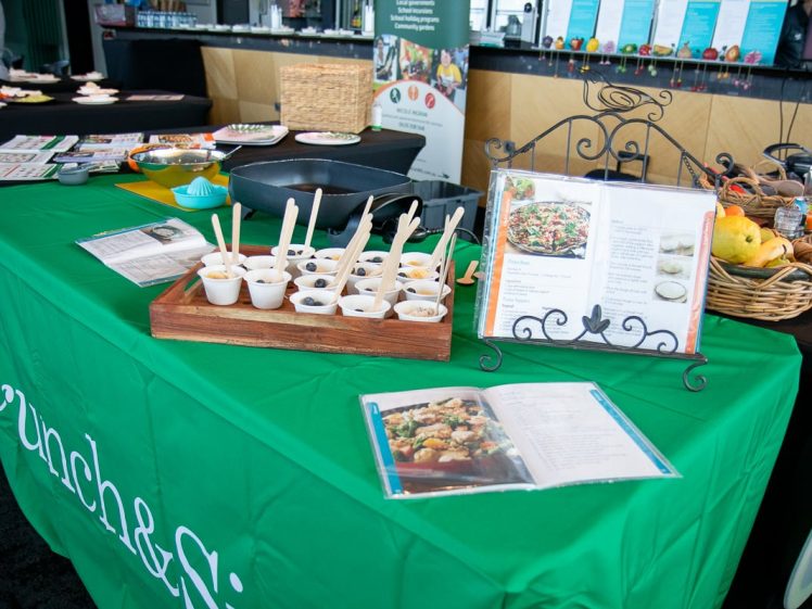 Image of A green Crunch & Sip table displays a recipe book, tasting samples in small white cups with spoons, a pan, fresh fruit in a basket, and informational leaflets at an event.