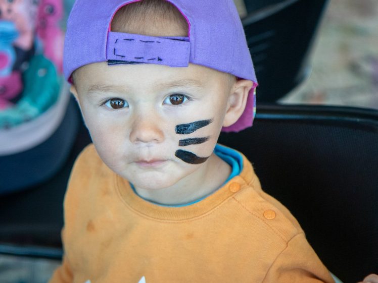 Image of A young child wearing a purple cap back to front and an orange shirt sits on a chair with three black face paint stripes on their cheek, looking at the camera.