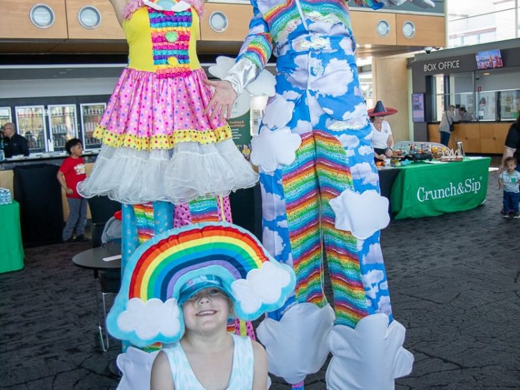 Image of Two performers on stilts dressed in colourful, whimsical cloud- and rainbow-themed costumes pose and smile indoors, while a child in a rainbow hat and blue dress stands happily in front of them.