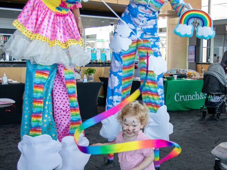 Image of A child with face paint plays with a rainbow ribbon in front of two performers on stilts dressed in colourful, whimsical costumes with clouds and rainbows. The scene is indoors at an event or festival.