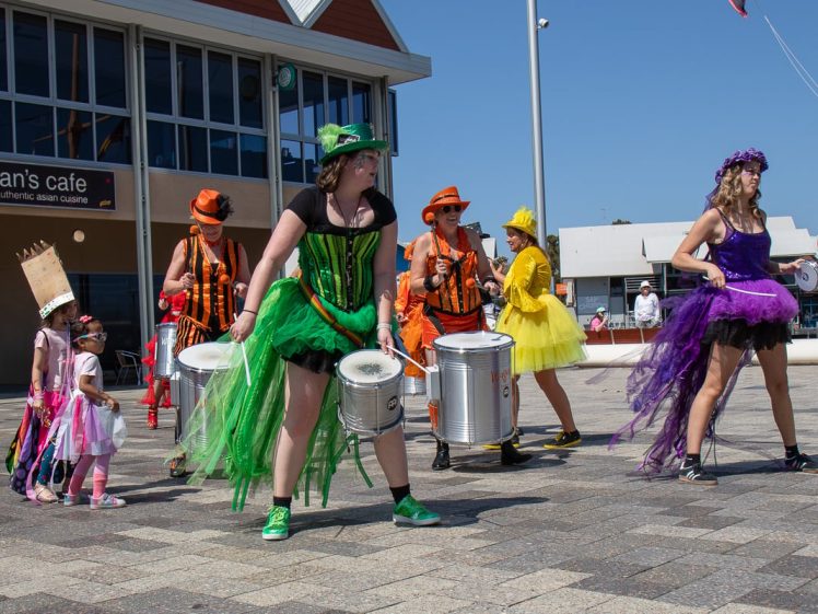 Image of A group of people in colourful costumes play drums outside on a sunny day. Two children in fancy dress stand nearby, watching. The scene takes place in a public square with shops and cafés in the background.