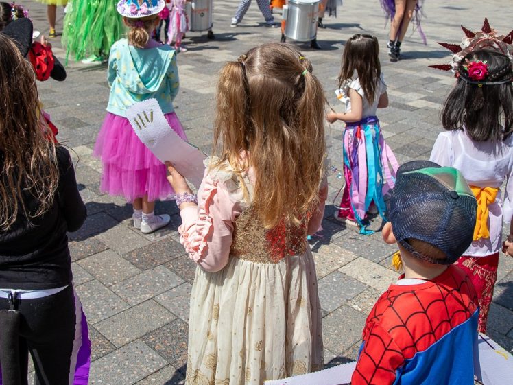 Image of Children in colourful costumes, including a superhero and princess, watch a lively outdoor performance by people playing drums. The event takes place on a paved area with buildings in the background.