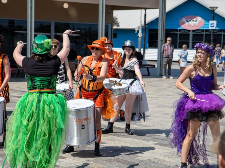 Image of A lively group of performers in colourful, whimsical costumes play drums outdoors. They wear bright skirts, hats, and accessories, and appear to be part of a parade or festival, entertaining a public audience in a sunny plaza.