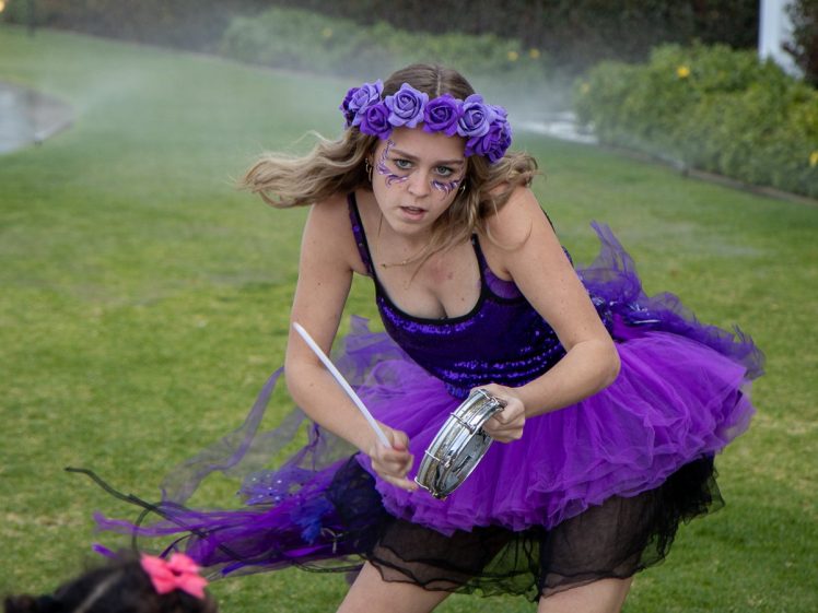 Image of A young woman in a purple tutu and floral crown plays a tambourine outdoors, with intense expression and face paint, as water sprays in the background.