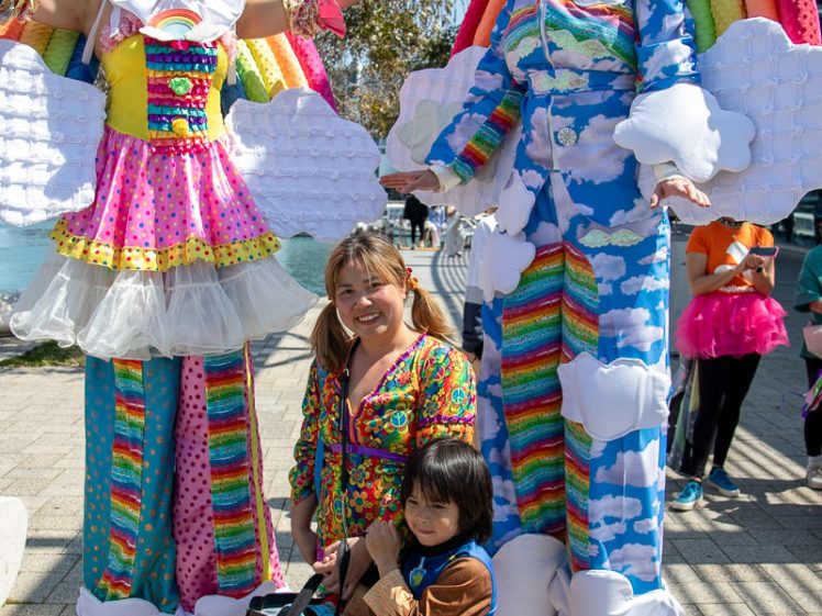 Image of Two adults on stilts wear colourful rainbow and cloud costumes, standing on either side of a smiling woman and child in fancy dress, outdoors on a sunny day. The group appears happy and festive.