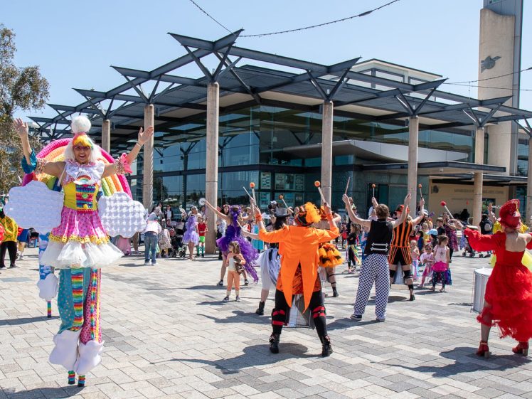 Image of A lively outdoor event with performers in colourful costumes, including a person on stilts, entertaining a crowd in front of a modern building. People of all ages watch and participate.