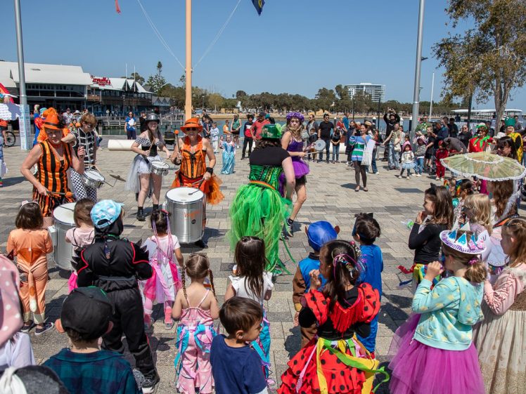 Image of A lively outdoor event with performers in colourful costumes playing drums and dancing, surrounded by children and adults dressed up, watching and enjoying the festivities in a public square.