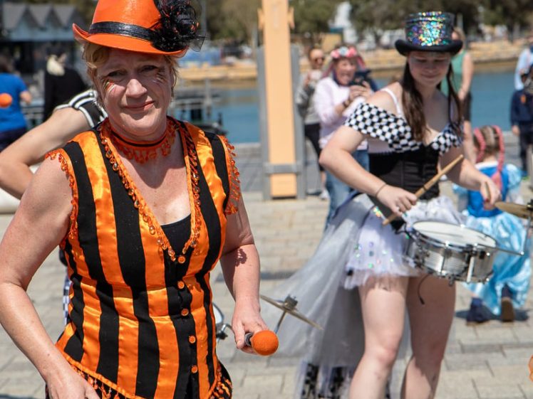 Image of A woman in an orange and black striped costume and hat plays a drum, whilst another woman in a chequered outfit and sparkly hat also drums. They perform outdoors near water with people watching in the background.