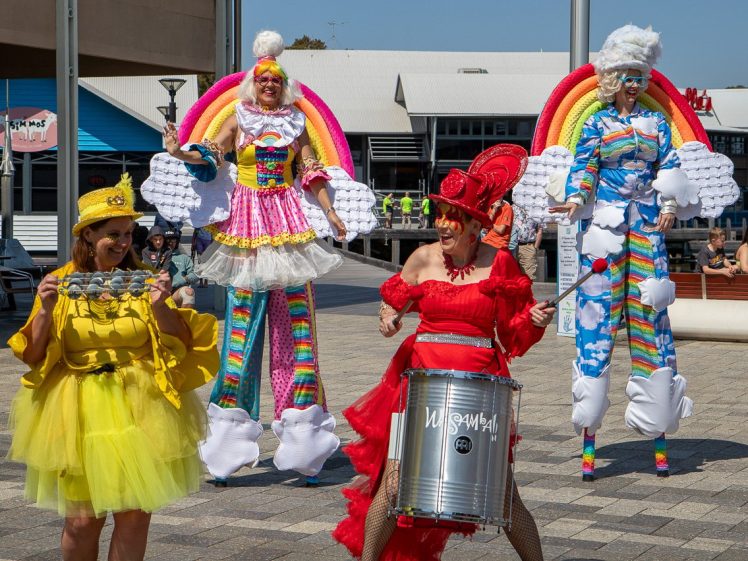 Image of Four performers in colourful costumes stand outdoors. Two on stilts wear rainbow outfits with white clouds, while the others wear bright yellow and red dresses; one holds a drum and the other holds an object, entertaining a crowd.