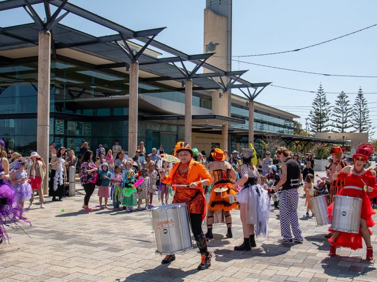 Image of A lively street performance with people in colourful costumes playing drums and dancing, entertaining a crowd gathered outside a modern building on a sunny day.