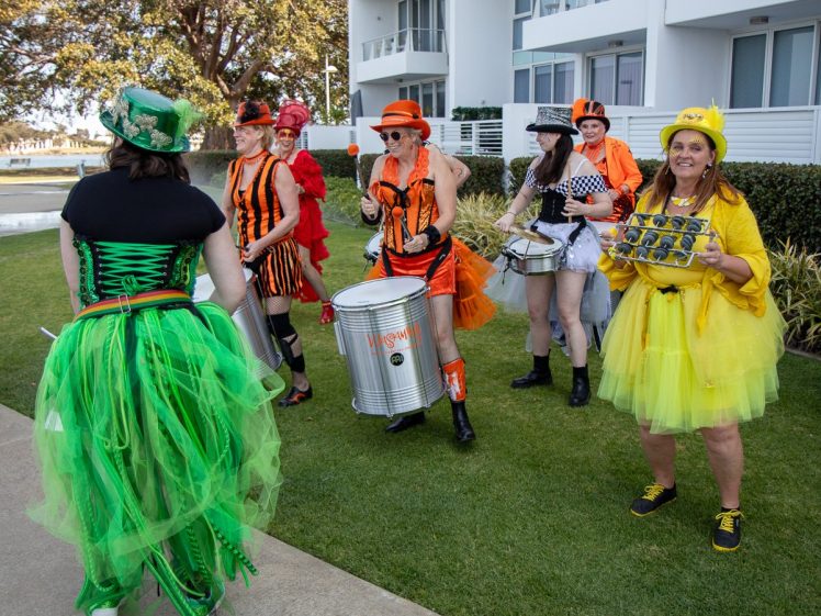 Image of A group of people in bright, colourful costumes play drums and percussion instruments outside a modern white building. One person wears a green dress, and another is in a yellow tutu holding bells. The group appears lively and festive.