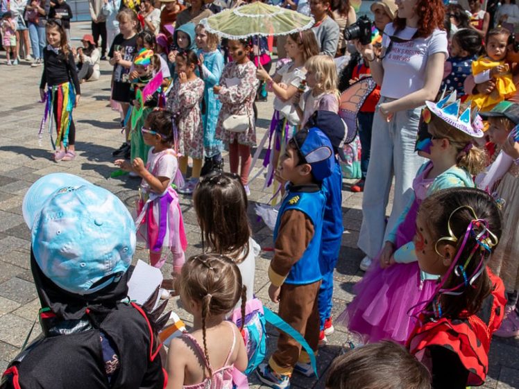 Image of A group of children in colourful costumes gather outdoors at an event. Adults are standing nearby, and trees and modern buildings are visible in the background under a sunny sky.