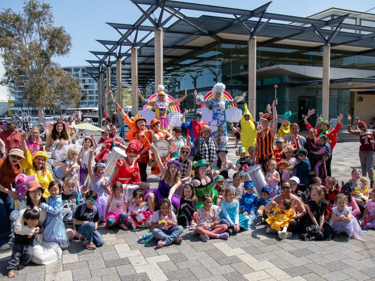 Image of A large group of children and adults in colourful costumes pose together outside a modern building on a sunny day. The crowd is smiling, waving, and some are dressed as clowns, superheroes, and animals.