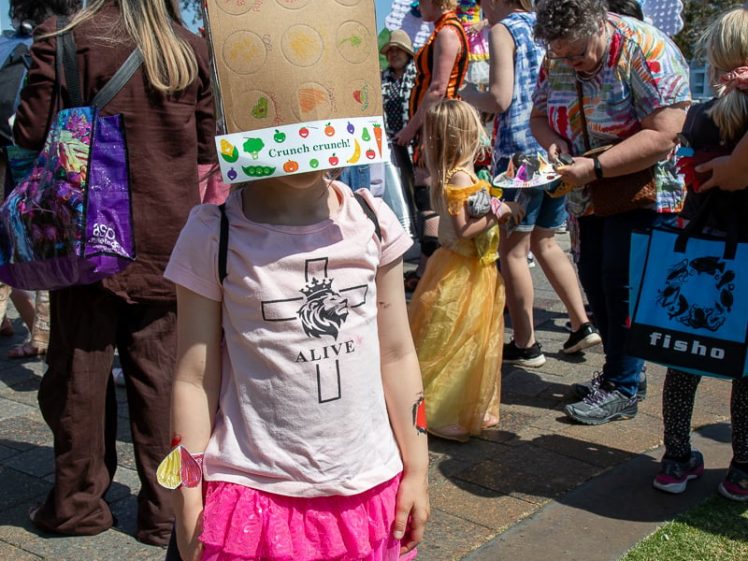 Image of A child stands outdoors wearing a decorated cardboard box as a hat, with a pink skirt and patterned tights, among a crowd of people in colourful costumes at a lively event.