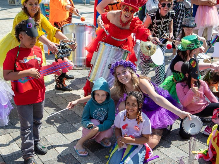 Image of A lively outdoor parade scene with adults and children in colourful costumes playing musical instruments and smiling. Some wear tutus, hats, and face paint, creating a joyful and festive atmosphere.