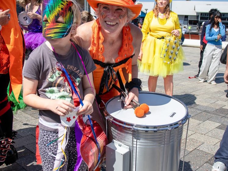 Image of A smiling adult in an orange costume and hat poses with a child holding a rainbow mask in front of a drum, surrounded by people in colourful outfits at an outdoor event.