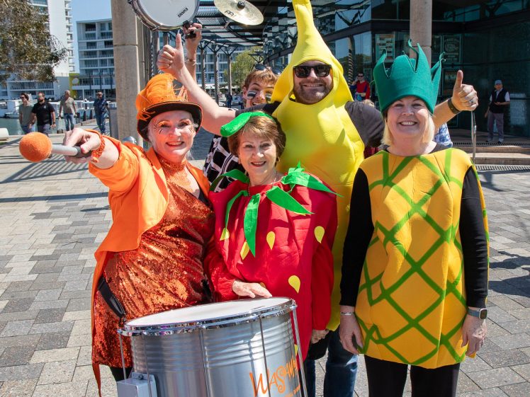 Image of Five adults dressed in bright fruit costumes—including a strawberry, banana, and pineapple—pose smiling outside by a drum. One person holds drumsticks, and another points playfully at the camera. It’s a sunny day.