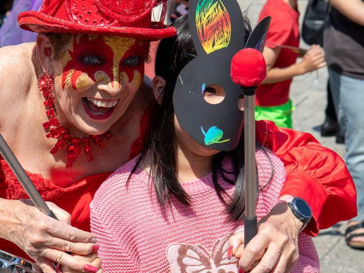 Image of A person in an elaborate red costume and headdress poses, smiling, with a child wearing a pink jumper and holding a decorated black rabbit mask over their face at an outdoor event. Other people are in the background.