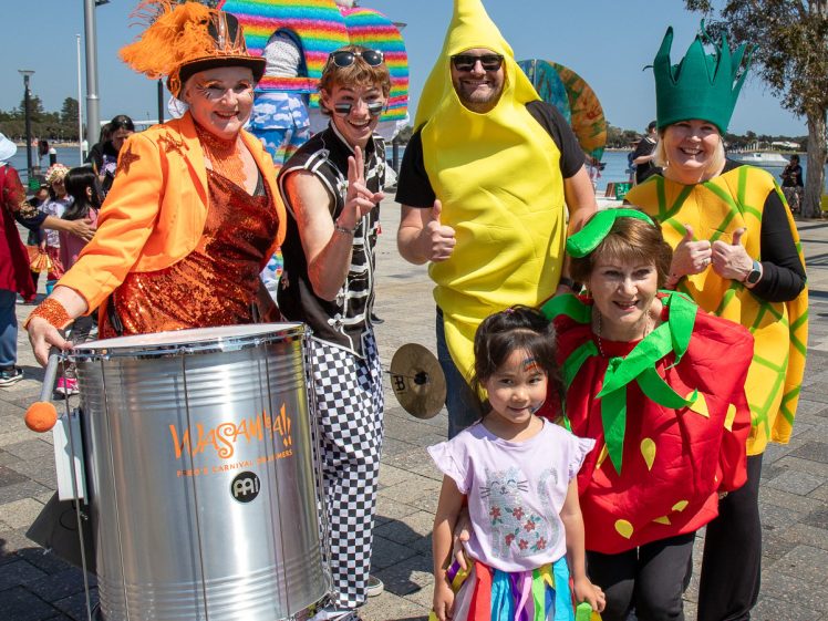 Image of A group of adults in colourful fruit and clown costumes smile and pose with a young girl at an outdoor event. Trees and other festival-goers are visible in the sunny background.