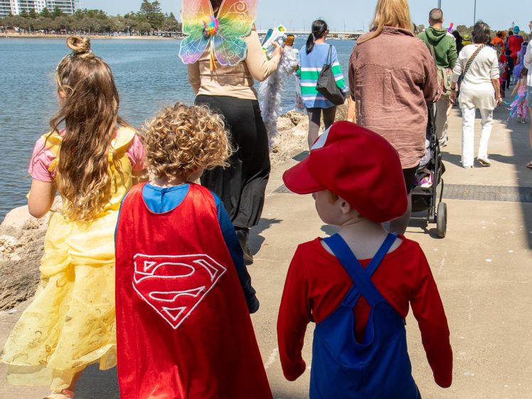 Image of Children dressed in colourful costumes, including Superman and Mario, walk along a waterfront path with adults and other children, some in fairy wings, during a sunny outdoor event.